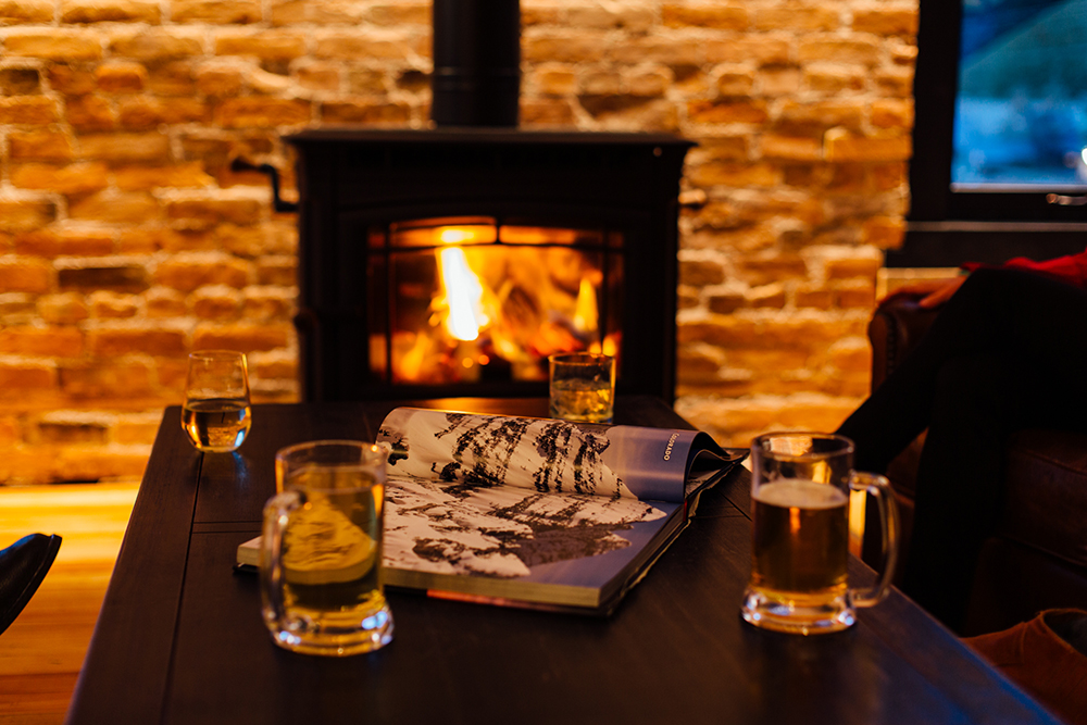 Image of a fire aglow in the wood buring stove casting a warm glow upon the coffee table adorned with an open book and some glass mugs of beer.